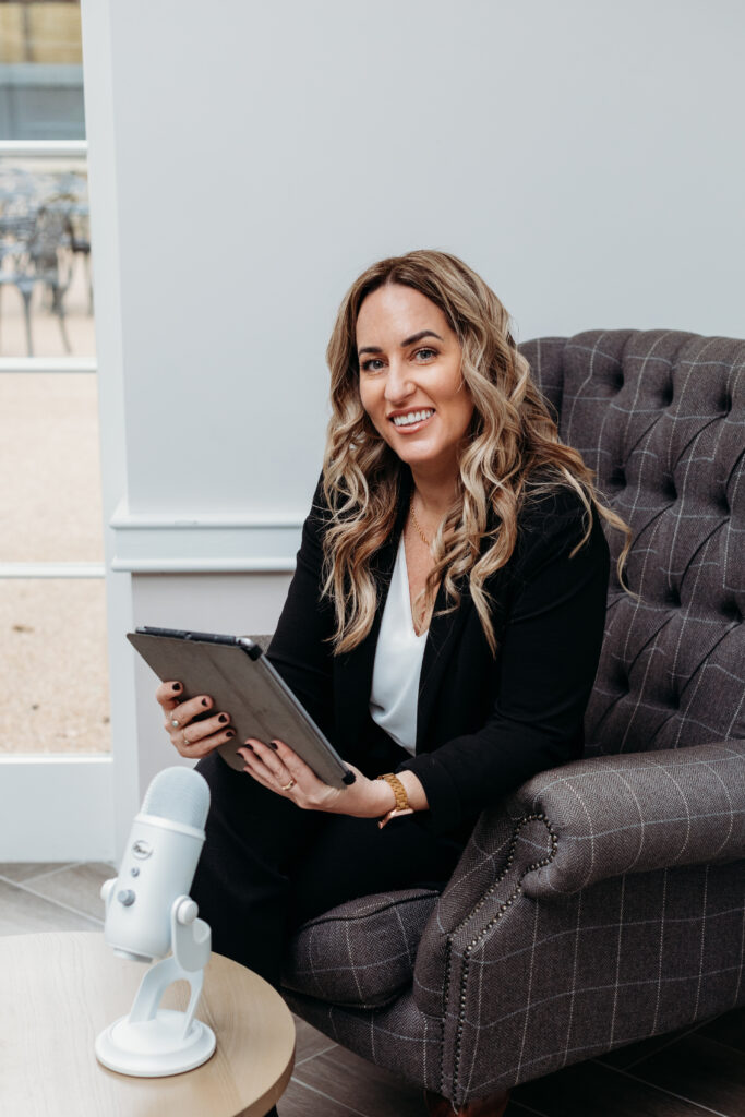 Gemma Heard talks about: Money mindset in business as she sits in a chair with an iPad in front of a microphone smiling at the camera.