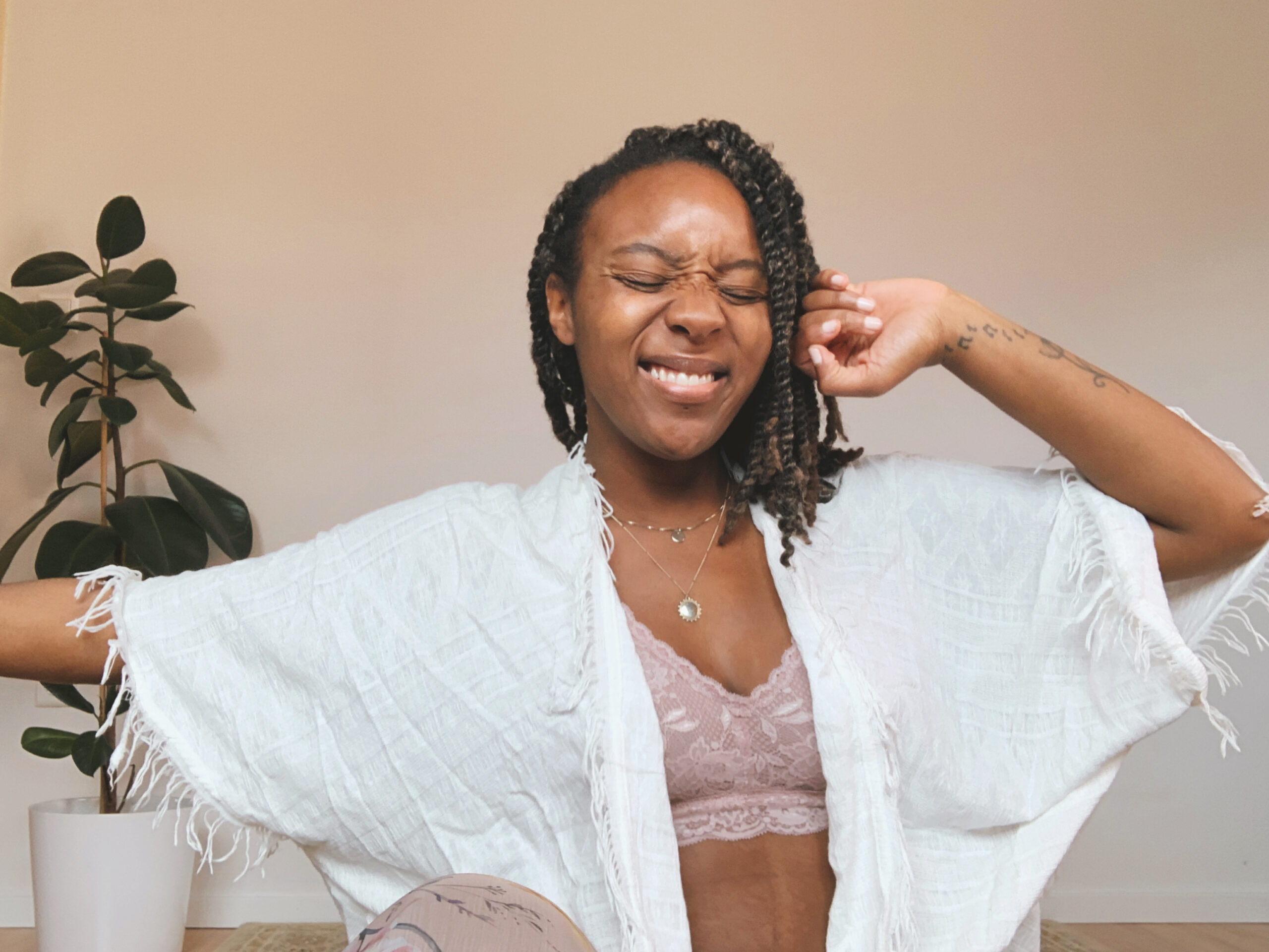 Natalie Martin, Menstrual cycle guru and black woman. Sits smiling with her eyes shut and arms outstretched She wears a white oversized shirt and bralette.
